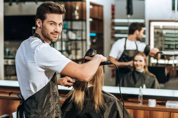 handsome young hairstylist smiling at camera while drying hair to beautiful young woman in beauty salon