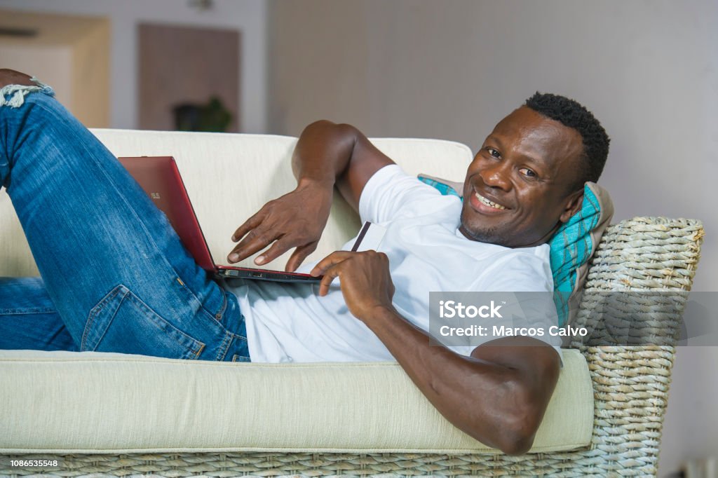 lifestyle portrait of young happy and attractive black afro American man using credit card and laptop computer relaxed and cheerful at living room sofa couch internet banking and online shopping 30-39 Years Stock Photo