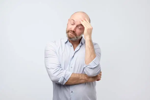 Portrait of tired and annoyed handsome european man iwith beard, leaning head on fist and breathing out from boredom, standing unhappy over gray wall