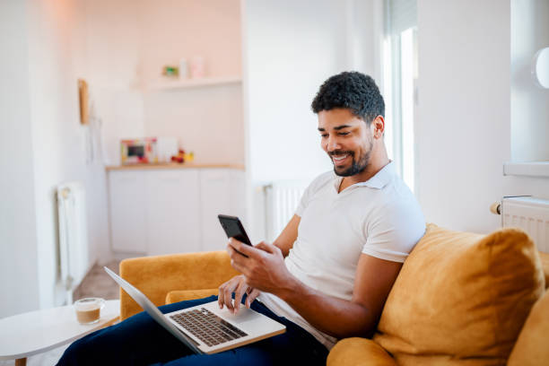 istock Smiling african american man working at home while sitting on the sofa. Using mobile phone and laptop. 1086603266