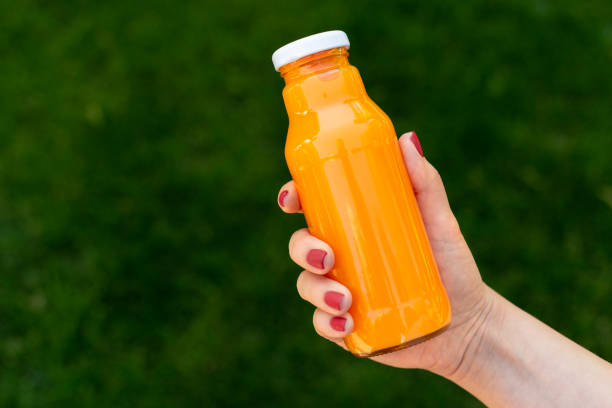 Vitamin concept, glass of fresh juice. Small glass bottle of fresh detox orange juice in woman's hand infront of green grass summer background. Vitamin concept. energy drink photos stock pictures, royalty-free photos & images