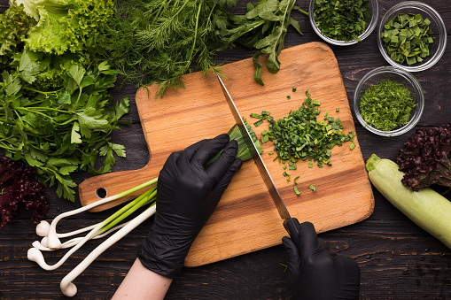 Chef hands in black gloves cutting spring onion on wooden board, kitchen table with fresh organic vegetables, top view