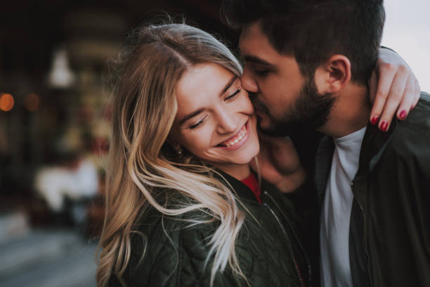 Beautiful young couple expressing their feelings on the street Precious moments of love. Close up portrait of handsome bearded guy kissing his girlfriend in cheek while she hugging him. Lady closing eyes with pleasure and smiling couple stock pictures, royalty-free photos & images