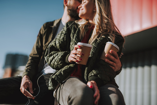 Charming girl holding cup of coffee while bearded man kissing and hugging her. Focus on female and male hands with beverages