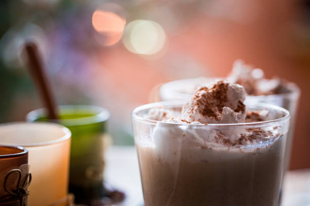 Close up color image depicting two glass tumblers full of freshly made eggnog, topped with whipped cream and dusted with cinnamon and nutmeg. The drinks are on a reflective glass surface and the defocused fairy lights of the Christmas tree are transformed into attractive glowing bokeh balls in the background. Room for copy space.