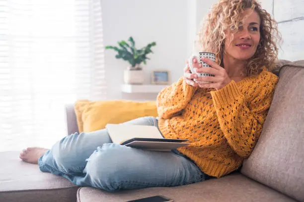 Photo of Beautiful 40 years old caucasian lady sit down on the sofa drinking tea and reading a book for afternoon indoor leisure activity at home - after work lifestyle for people concept