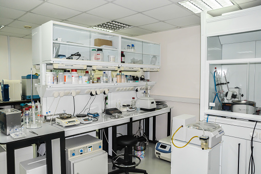 A young male Spanish scientist works carefully with a microscope at the laboratory using plastic gloves, protective eyewear and protective eyewear. He is holding the sample over the microscope and analyzing it carefully.