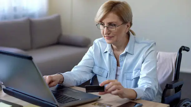 Senior woman entering credit card number on laptop, service for disabled people