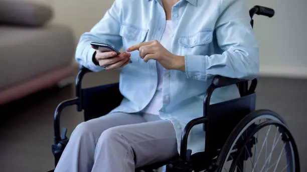 Female pensioner in wheelchair browsing internet on smartphone, communication