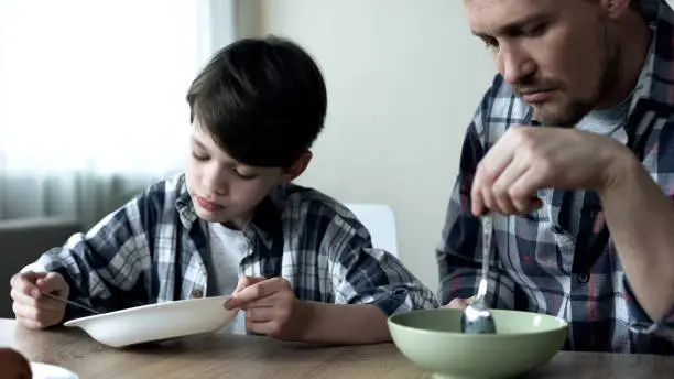 Serious single father and his son eating cornflakes in morning, poor breakfast