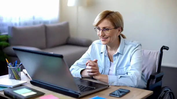 Positive elderly woman in wheelchair working on laptop, IT freelancer, online