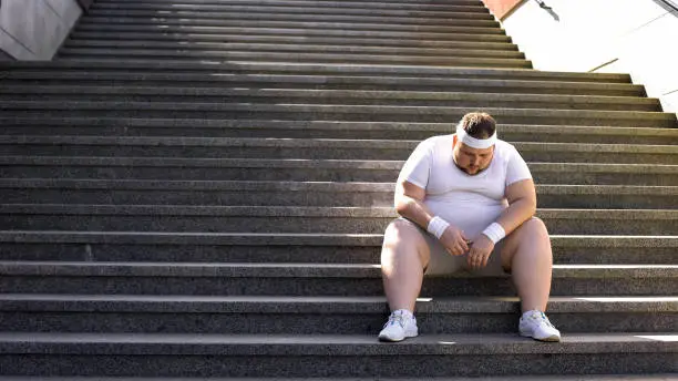 Photo of Upset man sitting alone on stairs, fighting with overweight and food addiction