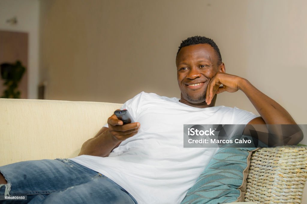 lifestyle portrait young attractive and happy black afro American man relaxed at home sofa couch enjoying watching television sports or movie smiling cheerful holding TV remote controller 30-39 Years Stock Photo