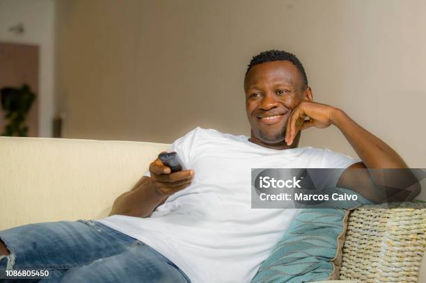 Lifestyle Portrait Young Attractive And Happy Black Afro American Man Relaxed At Home Sofa Couch Enjoying Watching Television Sports Or Movie Smiling Cheerful Holding Tv Remote Controller Stock Photo - Download Image Now