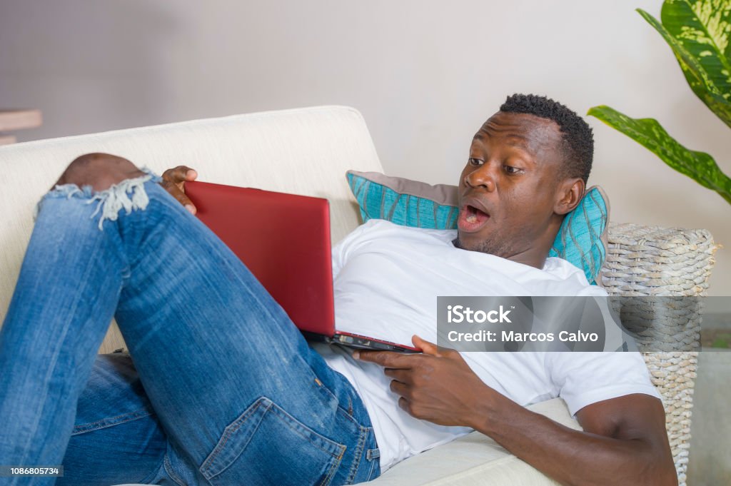 young excited and surprised young black African American man in disbelief and shock face expression networking with laptop computer lying at home living room couch with opened mouth 30-39 Years Stock Photo
