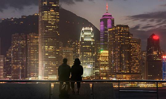 A young couple sitting on a waterfront barrier in the Kowloon district of Hong Kong, looking at the city lights of  Hong Kong Island across the sea channel
