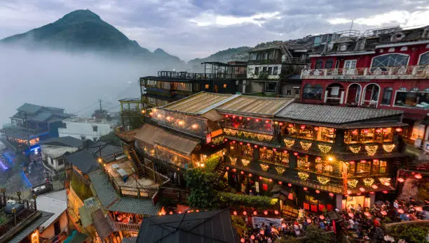 Slightly distorted wide angle shot of a teahouse lined with red lanterns with busy crowd in the lower right of the image and a mountain with a cloud inversion in the background.  Photographed in the historic mining town of Jiufen, Taiwan.