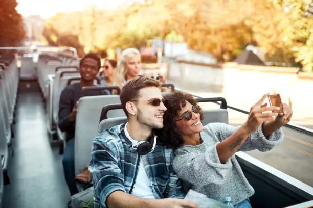 Group of tourists enjoying open top bus tour in the city. Focus on multi-ethnic couple making selfie.