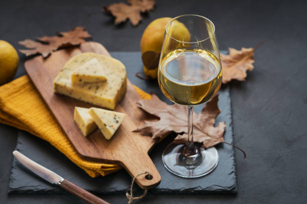 A glass of white wine served with cheese in a cutting board on dark background A glass of white wine was served with cheese in a cutting board on dark background. Autumn picnic with cheese, wine and dry leaves in rustic style. gouda cheese stock pictures, royalty-free photos & images
