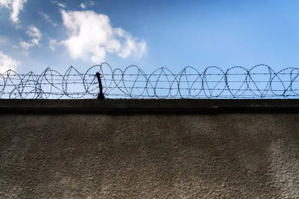 Photo of Barbed wire fence around prison walls, blue cloudy sky in background, security, crime or illegal immigration concept