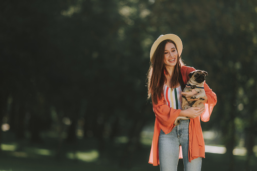 Waist up portrait of smiling woman in hat is holding pug puppy in her arms. Copy space on left side