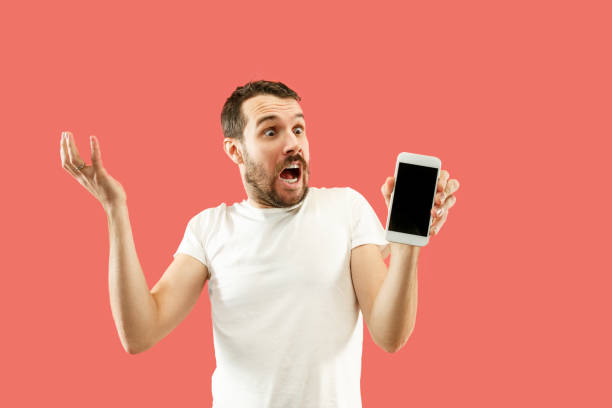 istock Young handsome man showing smartphone screen isolated on coral background in shock with a surprise face 1086940238
