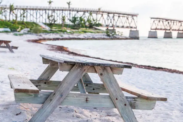 Sand beach, picnic table, bench at Bahia Honda state park, view on old, rust, rusty derelict rail road bridge, shore, coast, palm trees, sky at sunset, dusk, horizon, ocean, sea water