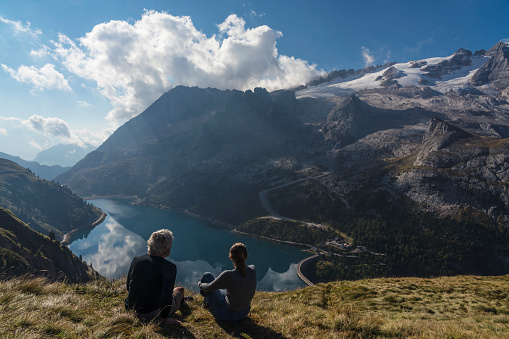 Senior couple hiking along hills opposite to the Marmolada in autumn. In distance lake Fedaia. Dolomites, Northern Italy.