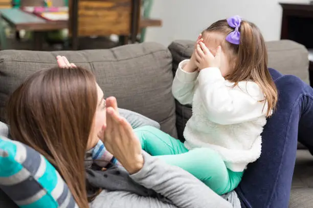 Joyful mid adult mother and cute daughter playing peekaboo on sofa at home