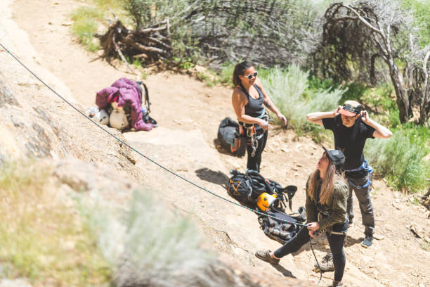 A millennial-age woman belays a climber. She is carefully holding the rope and watching her friend climb. Two others are standing close by putting on climbing harnesses.