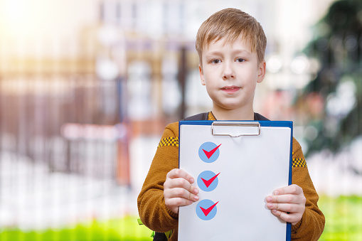 A schoolboy is standing at educational establishment blurred background and showing a plane table with a list of done tasks.