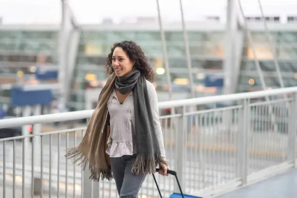 A young ethnic woman traveling solo pulls her suitcase through the airport. She is walking across an open-air skybridge. The terminal is in the background.