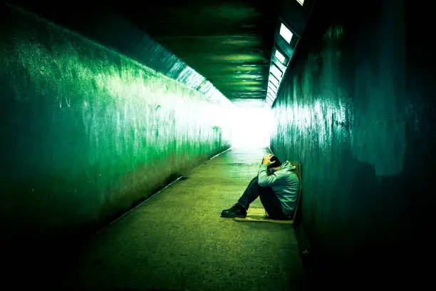 Color image depicting a young homeless man sitting alone in a cold, dark subway tunnel. The man is clearly sad, and has an unkempt beard. He sits with his back to the wall of the tunnel while the diminishing perspective of the subway tunnel recedes into the distance.