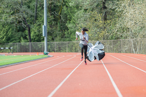 An African American athlete runs sprint drills on a stadium track. She is running with a parachute attached to her for resistance training.