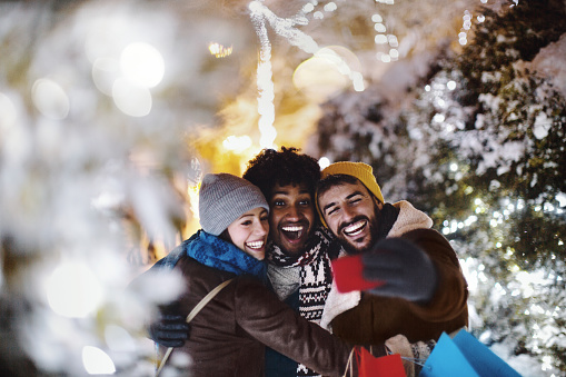 Young cheerful friends enjoying outdoors at winter time. They are taking selfie and smiling. They just finished shopping.