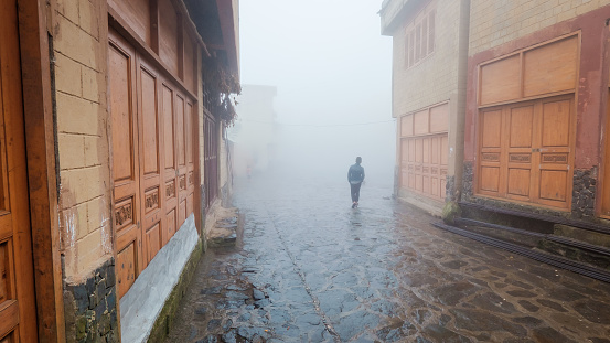 A lone person with hands in the pocket walking through a village street among a thick mist