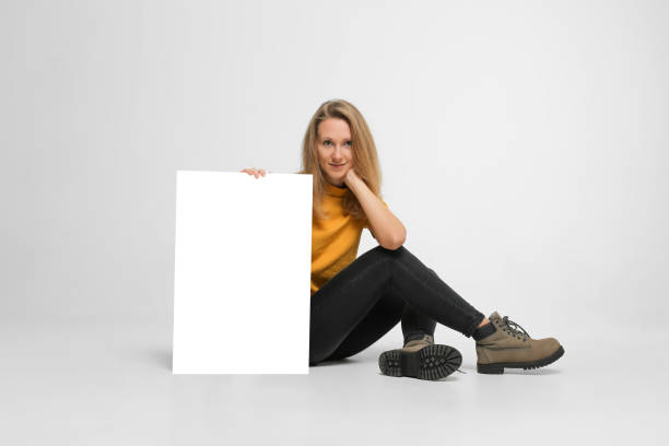 woman sitting with placard Young woman with blond hair, wearing casual outfit, boots, is sitting on white floor, holding white makeup poster, isolated on white background spokesmodel stock pictures, royalty-free photos & images