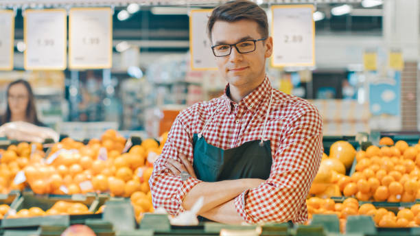 At the Supermarket: Portrait Of the Handsome Stock Clerk Wearing Apron, Arranging Organic Fruits and Vegetables, He Smiles and Crosses Arms. Friendly, Efficient Worker at the Store. At the Supermarket: Portrait Of the Handsome Stock Clerk Wearing Apron, Arranging Organic Fruits and Vegetables, He Smiles and Crosses Arms. Friendly, Efficient Worker at the Store. slow motion face stock pictures, royalty-free photos & images