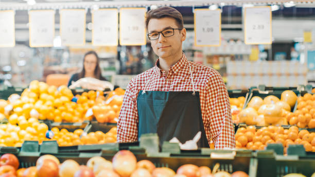 At the Supermarket: Portrait Of the Handsome Stock Clerk Wearing Apron, Arranging Organic Fruits and Vegetables, He Smiles into Camera. Friendly, Efficient Worker at the Store. At the Supermarket: Portrait Of the Handsome Stock Clerk Wearing Apron, Arranging Organic Fruits and Vegetables, He Smiles into Camera. Friendly, Efficient Worker at the Store. slow motion face stock pictures, royalty-free photos & images