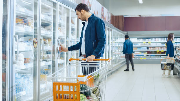 スーパーで: ハンサムな男は冷凍食品のコーナーで製品のショッピング カードやブラウザーをプッシュします。男は、ガラス扉の冷蔵庫、酪農製品を探しているに見えます。他の顧客は、バ� - 冷凍食品 ストックフォトと画像