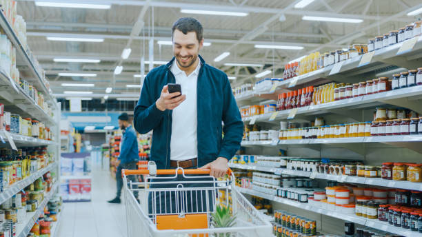istock At the Supermarket: Handsome Man Uses Smartphone, Smiles while Standing at the Canned Goods Section. Has Shopping Cart with Healthy Food Items Inside. Other Customers Walking in Background. 1087219642