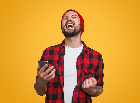 Handsome excited man in red beanie holding mobile phone and showing yes gesture with fist up standing on bright yellow studio background