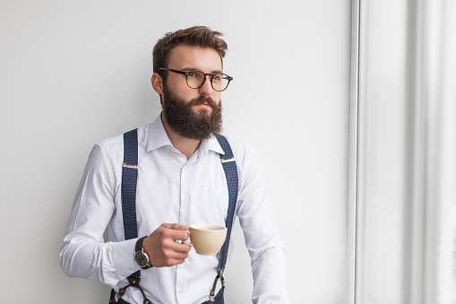 Serious bearded man in white shirt with suspenders holding cup of coffee and looking away in thoughts on white background