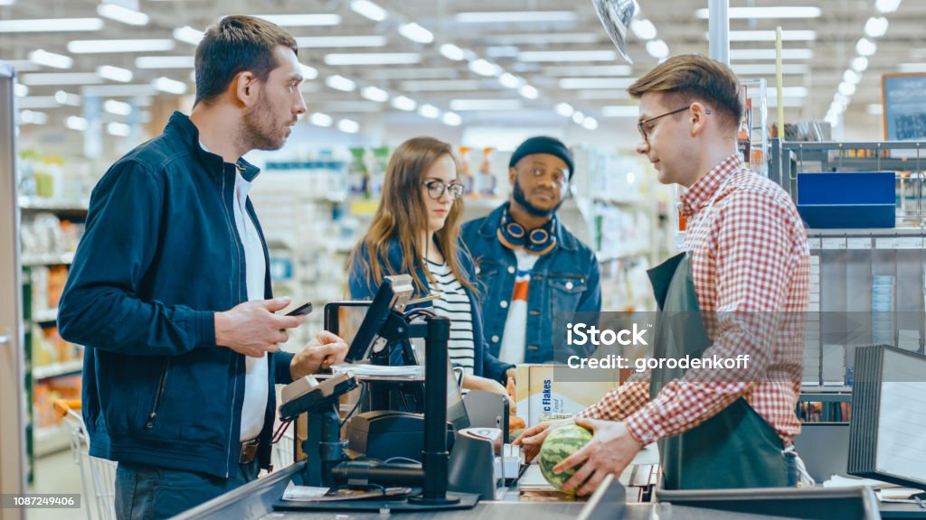 At the Supermarket: Checkout Counter Customer Pays with Smartphone for His Items. Big Shopping Mall with Friendly Cashier, Small Lines and Modern Wireless Paying Terminal System. Supermarket Stock Photo