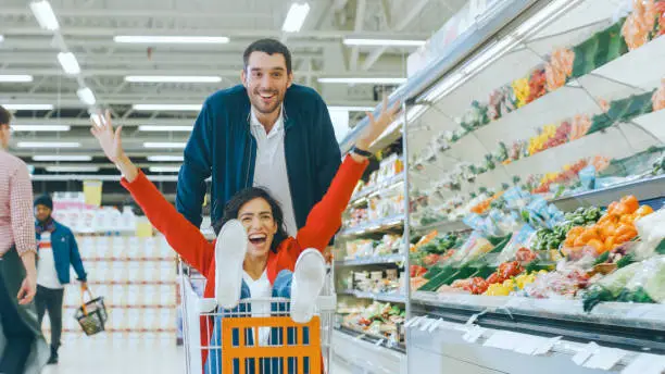 Photo of At the Supermarket: Man Pushes Shopping Cart with Woman Sitting in it, Happy Couple Has Fun Racing in a Trolley through the Fresh Produce Section of the Store. People Walking By.