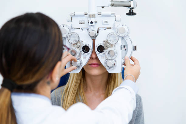 jeune femme assise sur une chaise avec beau opticien permanent tout en faisant un examen de la clinique d’ophtalmologie. - instrument optique photos et images de collection