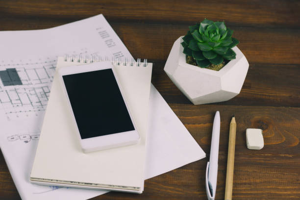 workplace with drawing plan, notepad, phone with blank black screen, pen, pencil, eraser, and succulent in concrete pot on wooden brown board background