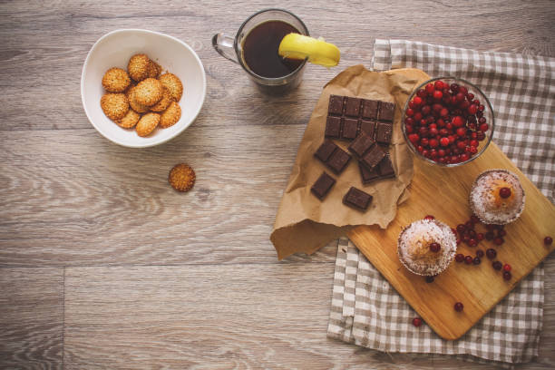 On a light wooden tabletop on a linen napkin napkin, there is a cutting board with two muffins, a broken chocolate bar and bright red berries in a small tree, next to a bowl with cookies.