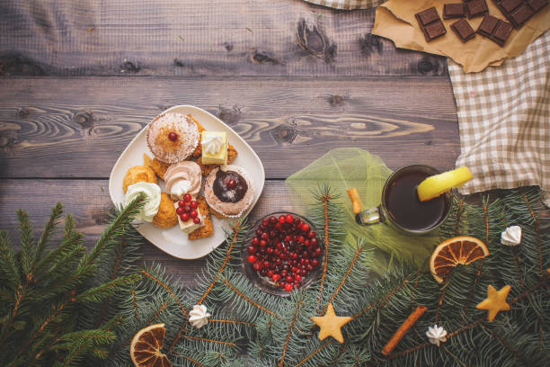 Happy New Year fir branches decorated with gingerbread stars, cinnamon sticks, dried orange slices and meringue peaks, bright cranberries, sweets in a dessert plate, a broken chocolate bar on kraft paper and a glass of tea with lemon on a linen napkin on a dark gray background. Festive pastry composition. feliz año nuevo ramas de abeto decoración con estrellas de pan de jengibre, canela, palillos, secado de rodajas de naranja y picos de merengue, arándanos rojos brillantes, dulces en un plato de postre, una barra de chocolate rota en papel kraft y un vas