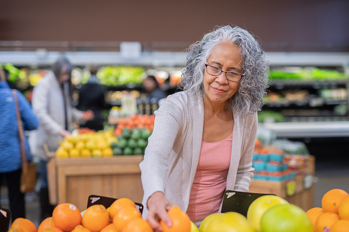 A cute older woman picks out fresh fruit at the supermarket. She is picking out lemons and inspecting each one carefully.